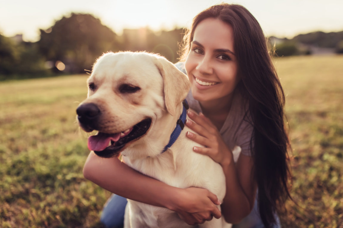 O tânără zâmbitoare îmbrățișând un câine Labrador Retriever auriu în lumina caldă a apusului, simbolizând armonia dintre proprietar și animalul de companie.